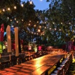 A wedding reception in a backyard in silicon valley. Market string lighting in a fanout pattern hangs above a long wood banquet table outside surrounded by tall trees. The long banquet table has a cobblestone pattern projected over it. The pattern is softly focused and covers the table and the ground. There are 4 strands of market lights visible in the image.