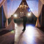 Photoshoot with fellow vendors showcasing custom chuppah with a scalloped pattern wash cast over white sheer drape. A couple stands towards the center of the large chuppah dancing. Two lights are centered on either side of the couple. Blue and white light glows around the room with a large golden chandelier with warm candlelit light glows from the candelabra bulbs.
