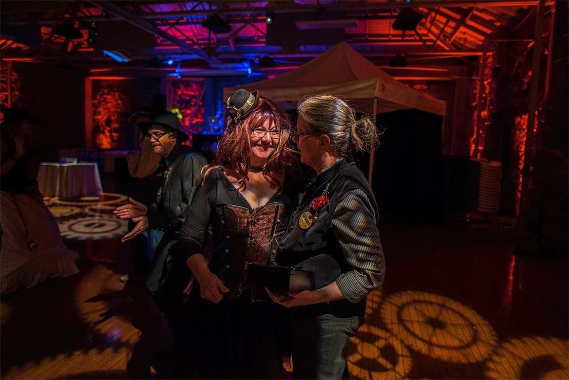 A woman with shoulder length red hair and a retro steampunk outfit is smiling while speaking with a woman with grayish silver hair, also in steampunk attire. They are surrounded by gears on the floor that have been projected in an orange color hue. Behind them are blue and magenta lights coming from a stage where the DJ has his setup. A soft light is cast to illuminate the woman's face with the red hair.