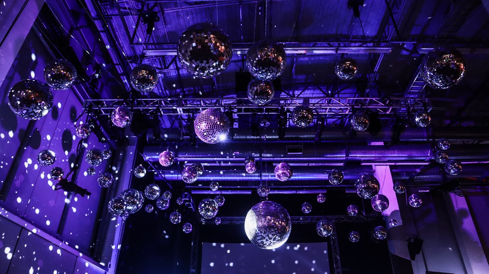 Verizon holiday party at the Yahoo Headquarters. Several mirror balls of varying sizes are hanging at different heights from an industrial style ceiling. The mirror balls are set in front of a purple color wash with some hints of magenta throughout.