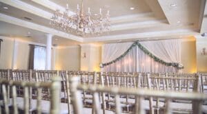 Wedding reception at Diablo Country Club showing a different angle with chiavari chairs blurred in the foreground and the ceremony area in focus. The backdrop to the ceremony is an ivory sheer drape wall with two sections of drape swagged from middle to side. Behind the wall of sheer drape are several edison pendants at varying heights producing a soft glow of light. The drape in the foreground has greenery attached to the edges. A large chandelier is in the middle of the room. Perimeter uplighting is seen in a soft amber near the drape.